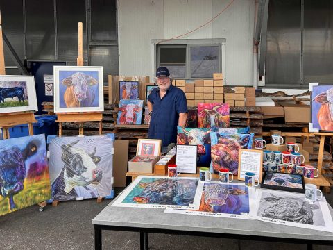 Der Künstler steht an seinem Verkaufsstand mit einer Vielzahl von Drucken, Tassen und Kissen seiner tierischen Werke. Die Produkte werden auf Staffeleien und Tischen präsentiert. Im Hintergrund befinden sich gestapelte Kartons mit Nachschub.