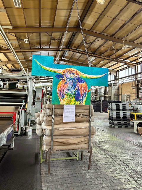 In einer großen Werkhalle hängt vor einem Lagergestell mit aufgerollten Materialien das bunte Triptychon eines Stieres an den Ketten eines Deckenkrans. Unter dem Gemälde befindet sich ein Informationsblatt zum Thema Welt ohne Fleisch. Die riesige Halle hat eine hohe Decke mit sichtbaren Metallträgern. Im Hintergrund sind industrielle Maschinen, Rohre und große Fenster zu sehen.