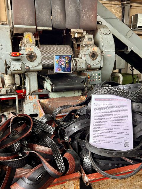 In einer Industrieumgebung hängt an einer Maschine ein buntes Gemälde eines Bullen. Vor der Maschine liegen die Reste von ausgehärteteten Gummistreifen. Ein Informationsblatt ist in der Nähe der Gummistreifen platziert. Die Umgebung sieht industriell und funktional aus, mit vielen mechanischen Teilen und Vorrichtungen.