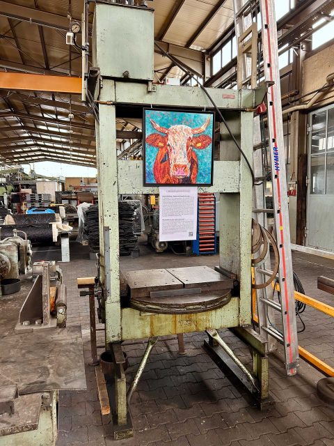 In einer Werkhalle hängt an einer Maschine von oben ein farbenfrohes Gemälde der Kuh Costa. Unterhalb des kleinformatigen Gemäldes ist ein Schild mit Informationen befestigt zum Thema Mutterkuhhaltung. Im Hintergrund sind industrielle Regale, Werkzeuge und Materialien zu sehen. Eine Leiter steht rechts neben der Maschine.