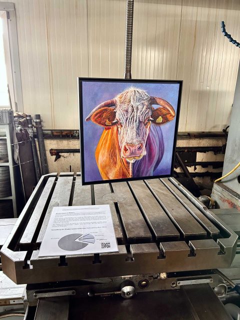 Das Porträt der Kuh Daisy mit blauem Hintergrund hängt an einer Industriemaschine. Vor dem Gemälde liegt das Infoblatt mit Infos über die Verteilung der Rinderrassen in Bayern. Im Hintergrund sind die helle Wand und einige Metallteile zu sehen.