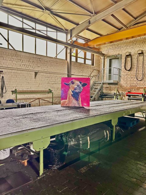In einer großen Industriehalle steht das farbenfrohe Gemälde eines Kamerunschafes auf einem breiten Förderband. Im Hintergrund sind weiße Backsteinwände, große Fenster und eine gelbe Kranbahn zu sehen. Eine Metalltreppe führt zu einer Tür.