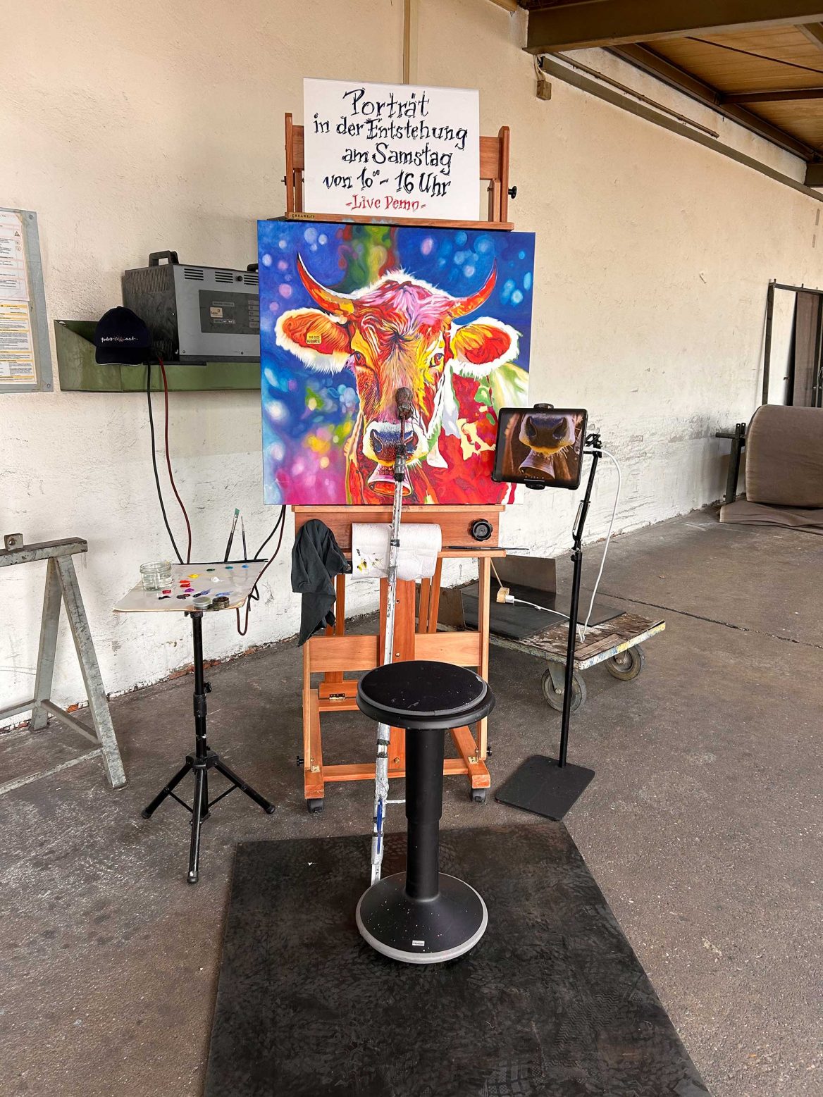 Das leuchtend bunte Kuhporträt von „Huberte” ist noch nicht beendet und steht auf einer Staffelei in einer hellen Industriehalle. Über dem Gemälde ist ein Schild befestigt, auf dem steht: "Porträt in der Entstehung am Samstag von 10-16 Uhr, Live Demo." Daneben befinden sich ein Tisch mit Malutensilien sowie ein Stuhl, der auf einer schwarzen Gummimatte steht. Im Hintergrund ist eine weiße Wand mit verschieden Industriegegenständen zu sehen.