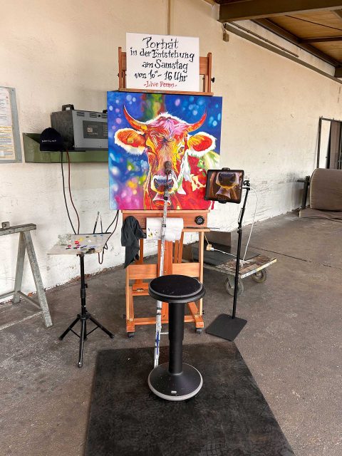 Das leuchtend bunte Kuhporträt von „Huberte” ist noch nicht beendet und steht auf einer Staffelei in einer hellen Industriehalle. Über dem Gemälde ist ein Schild befestigt, auf dem steht: "Porträt in der Entstehung am Samstag von 10-16 Uhr, Live Demo." Daneben befinden sich ein Tisch mit Malutensilien sowie ein Stuhl, der auf einer schwarzen Gummimatte steht. Im Hintergrund ist eine weiße Wand mit verschieden Industriegegenständen zu sehen.
