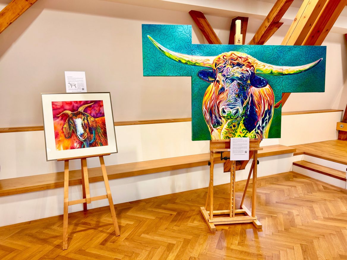 Zwei Gemälde von Tieren auf Staffeleien. Links das Aquarell-Porträt einer Ziege in kräftigen Farben, rechts ein großes, farbenfrohes Porträt eines Longhornbullen als Tripütychon in Öl. Bei beiden Gemälden hängen Infoschilder daneben.
