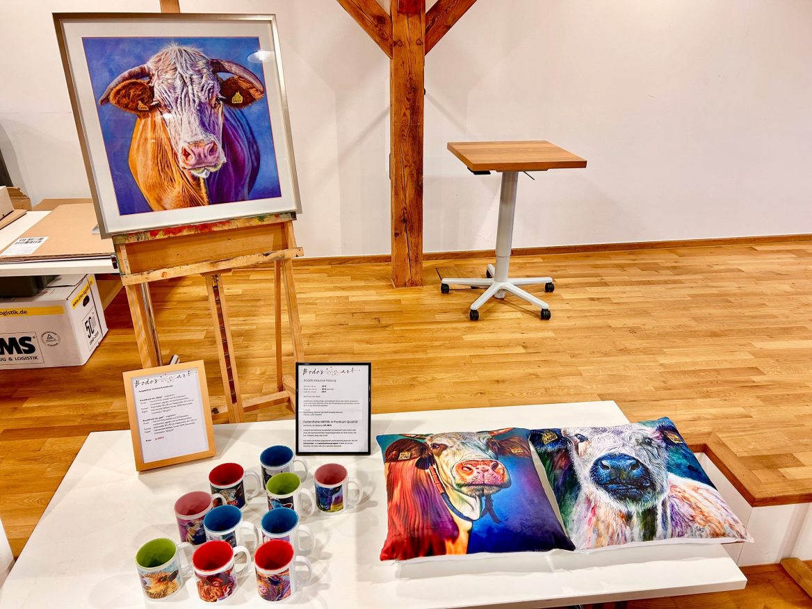 Auf einer Staffelei steht ein gerahmter, hochwertiger Kunstdruck des Porträtgemäldes von der Kuh Grace. Vor der Staffelei stehen auf einem Tisch Keramiktassen mit Gsedls Tierporträts und rechts daneben liegen zwei Kissen bedruckt mit den Motiven von Joni und Anastasia. Zwei Schilder mit Informationen und Preisen sind ebenfalls aufgestellt. Im Hintergrund ist ein kleiner Tisch sichtbar.