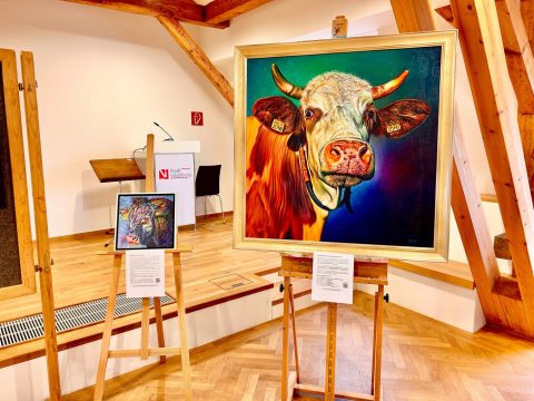 Zwei farbenfrohe Gemälde von Rindern sind jeweils auf einer Staffelei ausgestellt. Das größere Bild zeigt ein detailliertes Porträt der Kuh Joni. Links daneben das kleinere Gemälde eines Stierkopfes. Im Hintergrund ist ein Rednerpult mit der Aufschrift "Stadt Vilsbiburg" zu sehen.