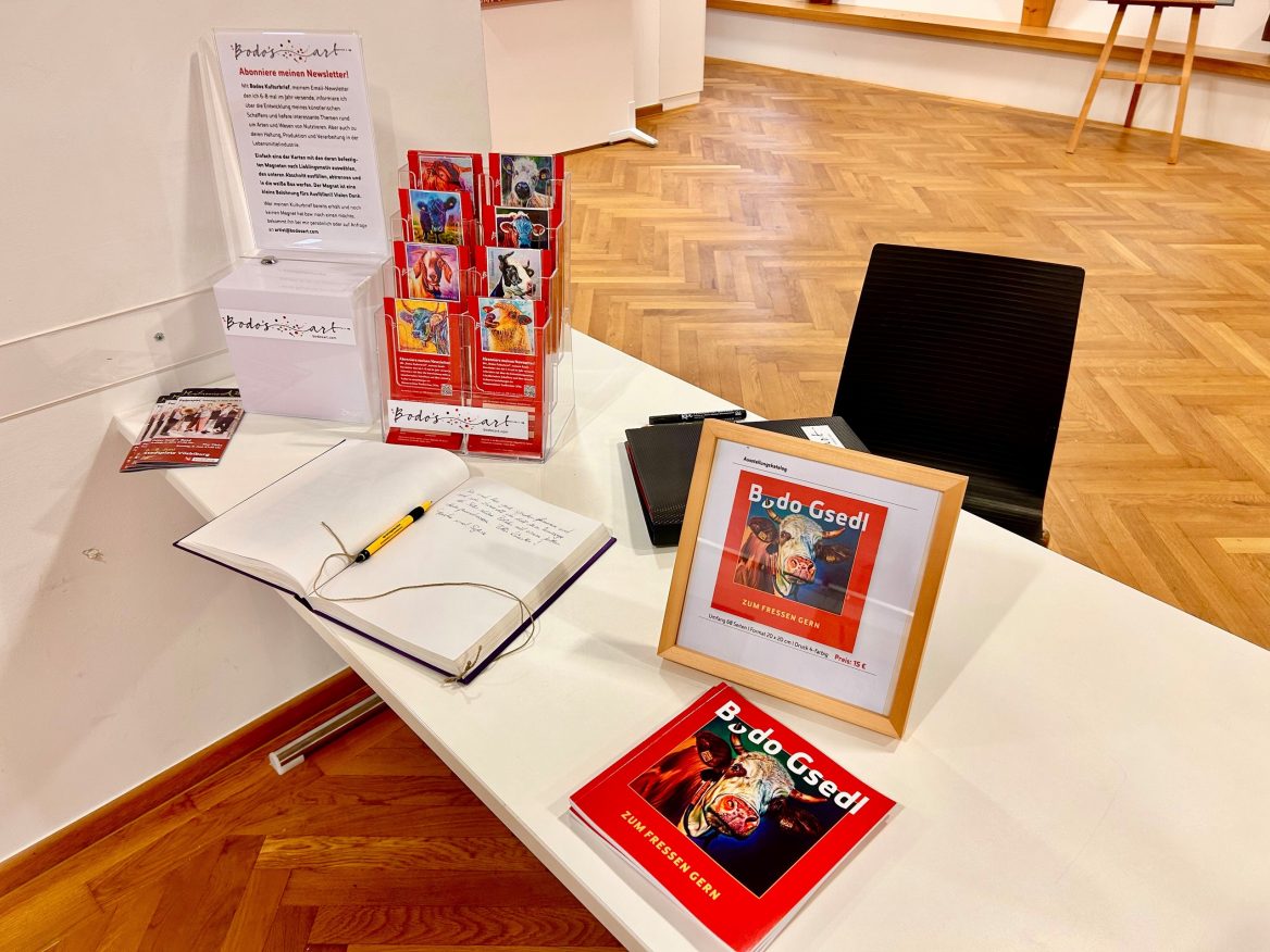 Ein Tisch mit Flyern, einem offenen Gästebuch mit Stift sowie dem Ausstellungskatalog des Künstlers Bodo Gsedl sowie ein gerahmtes Foto, das für den Kauf des Kataloges wirbt. Links daneben befindet sich ein Aufsteller, der zur Anmeldung für einen Newsletter einlädt. Im Hintergrund ist der helle Holzboden des Raumes sichtbar.