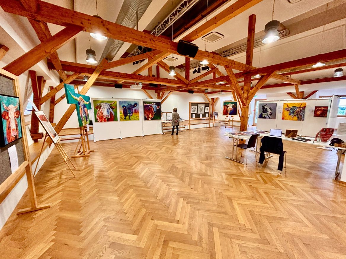 Ein geräumiger Galerieraum mit Holzboden und sichtbaren Holzbalken an der Decke. An den Wänden und auf Staffeleien sind bunte Tiergemälde ausgestellt. Eine Person betrachtet die Kunstwerke. Auf der rechten Seite steht ein Tisch mit einem Laptop und Stühlen. Der Raum ist hell beleuchtet.