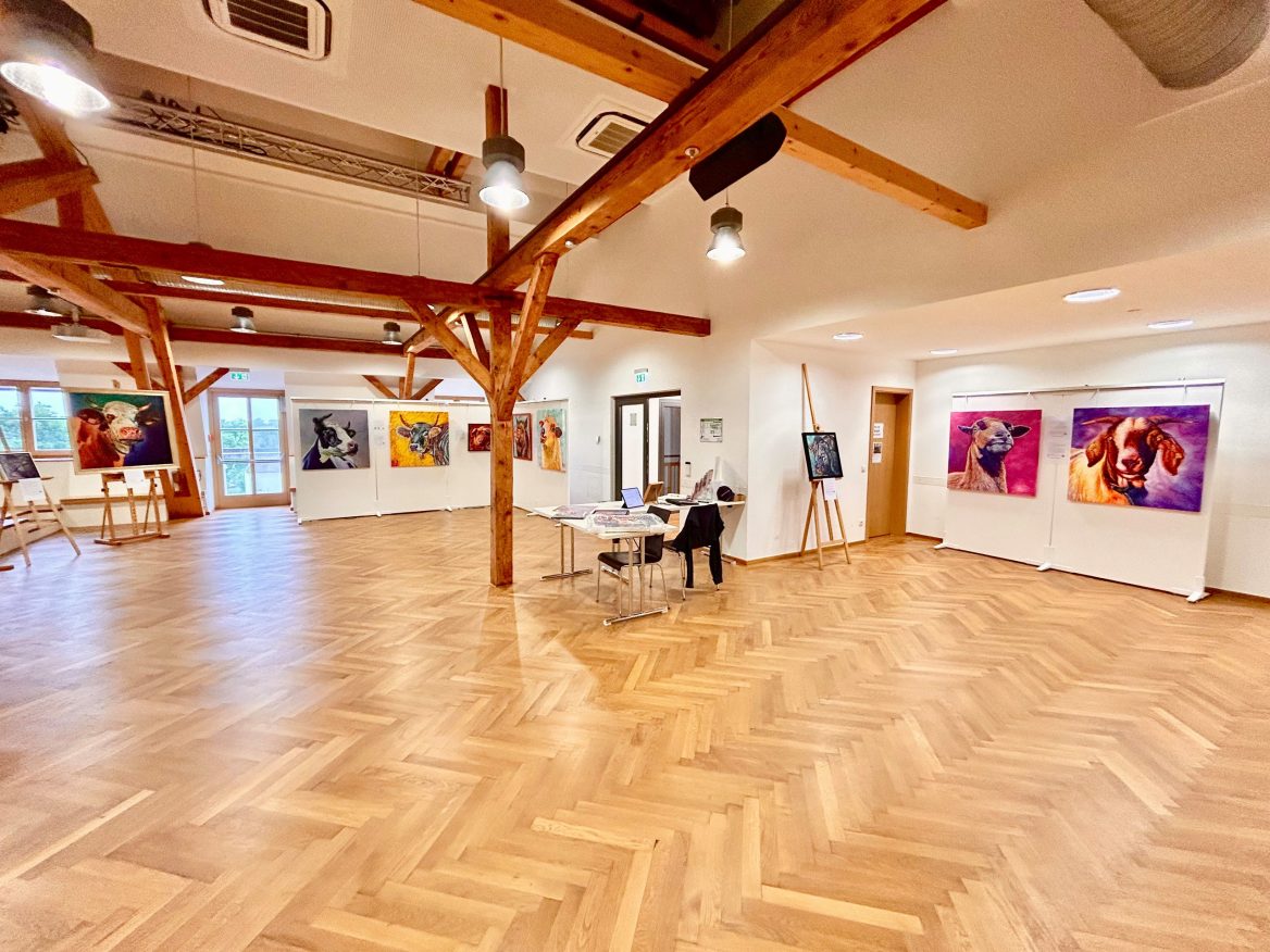Ein geräumiger, heller Galerieraum mit Holzboden und Holzbalken an der Decke. An den Wänden hängen bunte Gemälde von Nutztieren des Künstlers Bodo Gsedl. In der Mitte stehen ein Tisch und ein Stuhl. Der Raum hat große Fenster sowie moderne Beleuchtung.