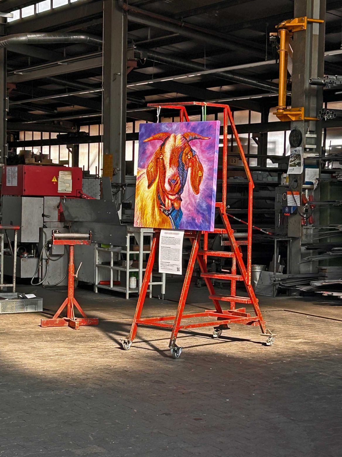 In einer Industriehalle steht ein leuchtend buntes Gemälde einer Ziege auf einer roten, mobilen Treppe. Das Gemälde ist mit Ketten an den oberen Sprossen befestigt, und darunter hängt ein Informationsblatt. Im Hintergrund sind Maschinen, Regale und Metallstrukturen zu sehen. Der Raum ist von Sonnenlicht durchflutet, das durch die Fenster an der Decke fällt.