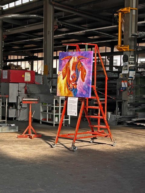 In einer Industriehalle steht ein leuchtend buntes Gemälde einer Ziege auf einer roten, mobilen Treppe. Das Gemälde ist mit Ketten an den oberen Sprossen befestigt, und darunter hängt ein Informationsblatt. Im Hintergrund sind Maschinen, Regale und Metallstrukturen zu sehen. Der Raum ist von Sonnenlicht durchflutet, das durch die Fenster an der Decke fällt.
