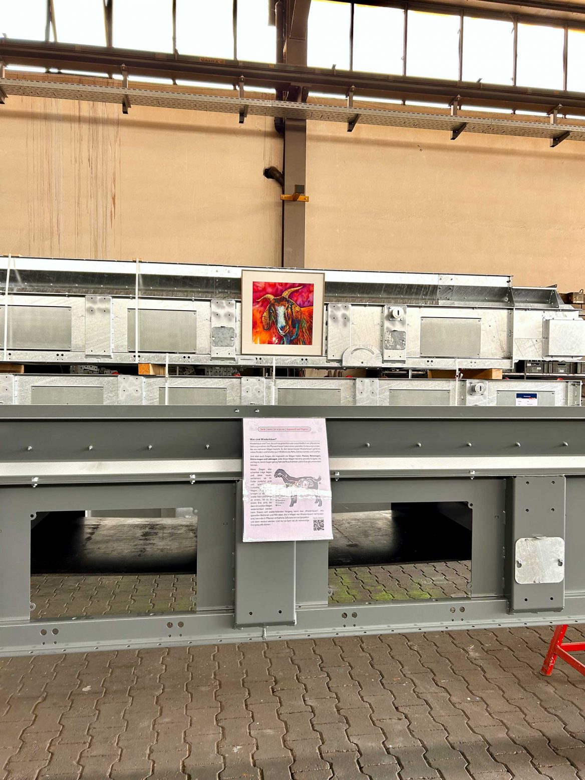 In einer Industriehalle hängt das bunte Aquarellgemälde des Ziegenbocks Jack an einem großen, metallischen Maschinenbauteil. Darunter hängt im Vordergrund ein Informationsblatt mit Text.