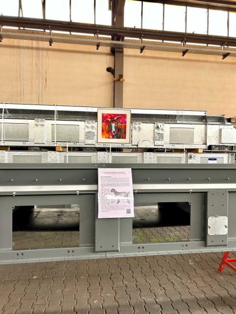 In einer Industriehalle hängt das bunte Aquarellgemälde des Ziegenbocks Jack an einem großen, metallischen Maschinenbauteil. Darunter hängt im Vordergrund ein Informationsblatt mit Text.