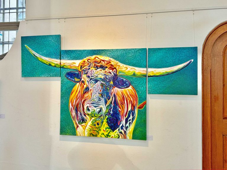 Ein farbenfrohes Triptychon-Gemälde zeigt einen Stier mit langen Hörnern. Der Stier ist in lebhaften Farben wie Orange, Gelb, Blau und Grün dargestellt. Der Hintergrund ist türkisfarben mit geschwungenen Mustern. Das Gemälde hängt an einer weißen Wand, neben einer hölzernen Türöffnung.