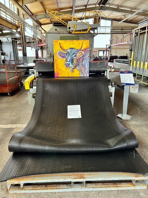 In einer Industriehalle steht ein buntes Gemälde einer Kuh auf einer schwarzem, strukturierten Gummimatte, das aus einer Maschine heraushängt. Vor dem Gemälde liegt ein Informationsblatt. Die Halle hat hohe Decken mit großen Fenstern, die viel Licht hereinlassen. Im Hintergrund sind verschiedene industrielle Strukturen und Ausrüstungen zu sehen.