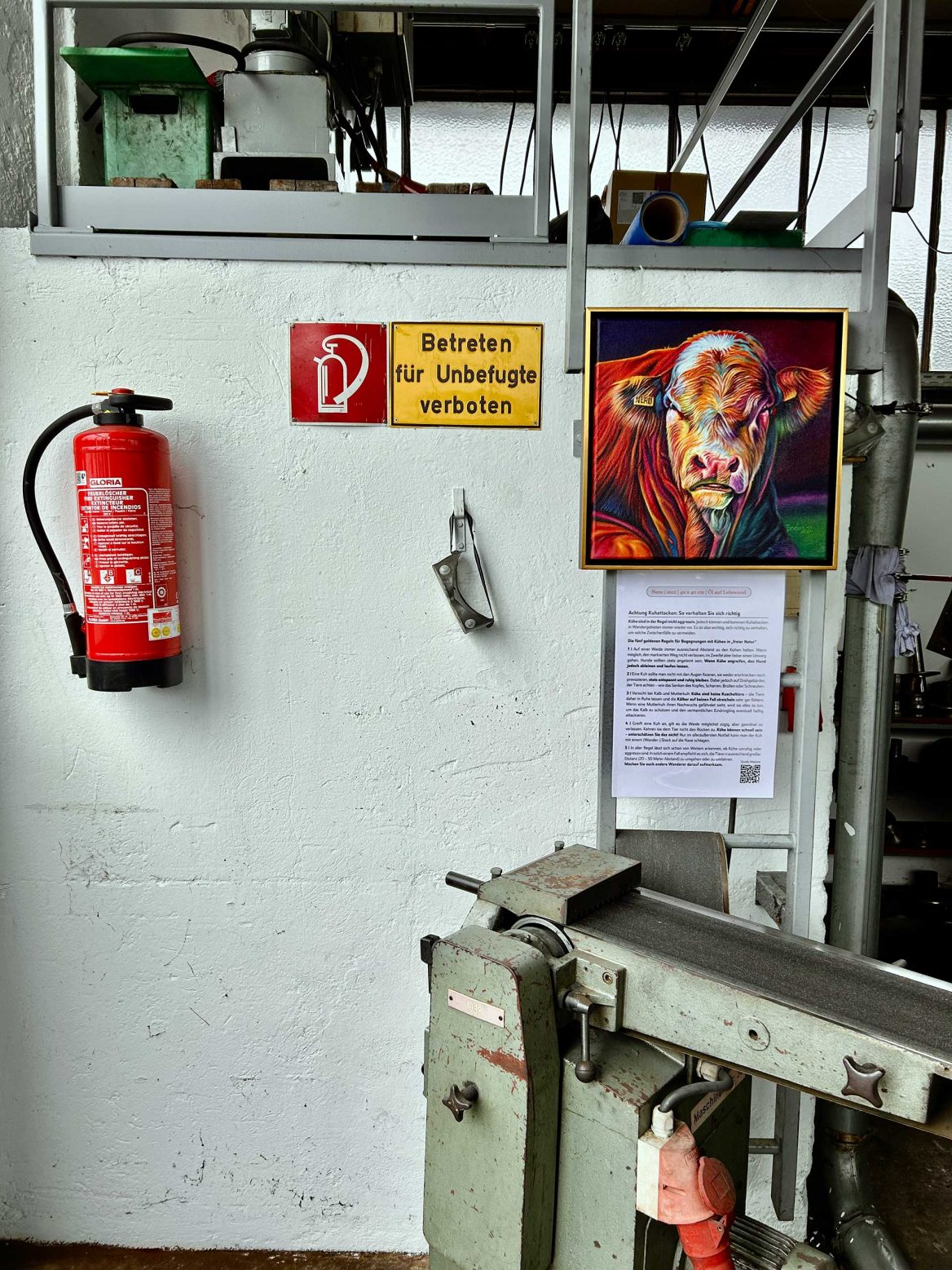 In einer Fertigungshalle hängt an einer weißen Wand ein buntes Gemälde eines Bullen. Daneben befindet sich ein Schild mit der Aufschrift „Betreten für Unbefugte verboten“ sowie ein Feuerlöscher. Unter dem Bild ist ein längerer Text befestigt. Im Vordergrund ist ein graues Metallgerät zu sehen, und im Hintergrund sind Regale mit verschiedenen Gegenständen.