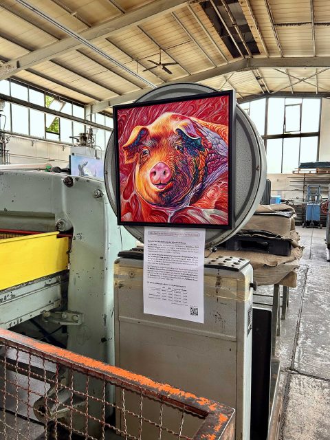 In einer Industriehalle hängt ein buntes Gemälde eines Schweins an der Rückseite einer großen Industriewaage. Darunter ist ein Infoblatt befestigt. DieHalle hat hohe Decken und große Fenster, durch die Tageslicht einfällt. Im Vordergrund ist ein Teil einer Transportkiste zu sehen, und im Hintergrund sind weitere industrielle Geräte und Strukturen erkennbar.