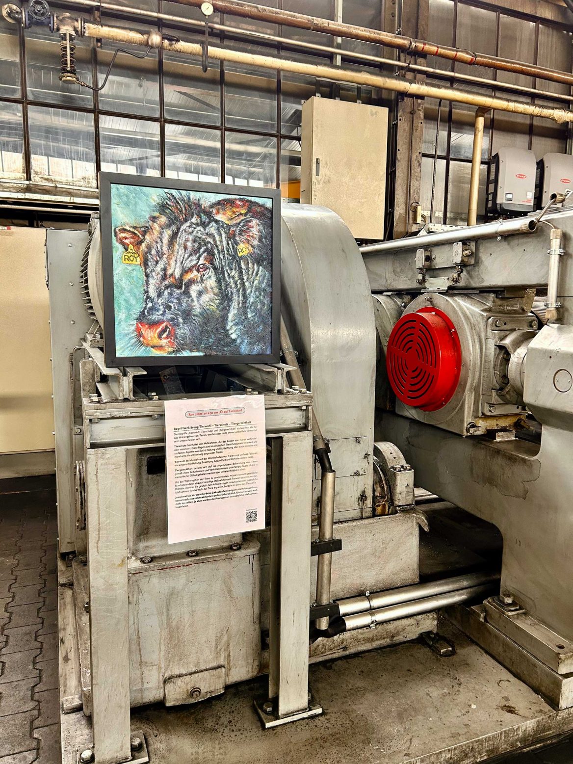 In einem industriellen Raum ist eine große graue Maschine zu sehen. Auf der Maschine ist ein farbiges Gemälde einer Kuh angebracht. Darunter befindet sich ein beschriftetes Blatt Papier mit Details. Im Hintergrund sind Rohre, große Fenster und weitere industrielle Elemente erkennbar. Die Maschine hat metallische Strukturen und ein rotes Bauteil ist deutlich sichtbar.