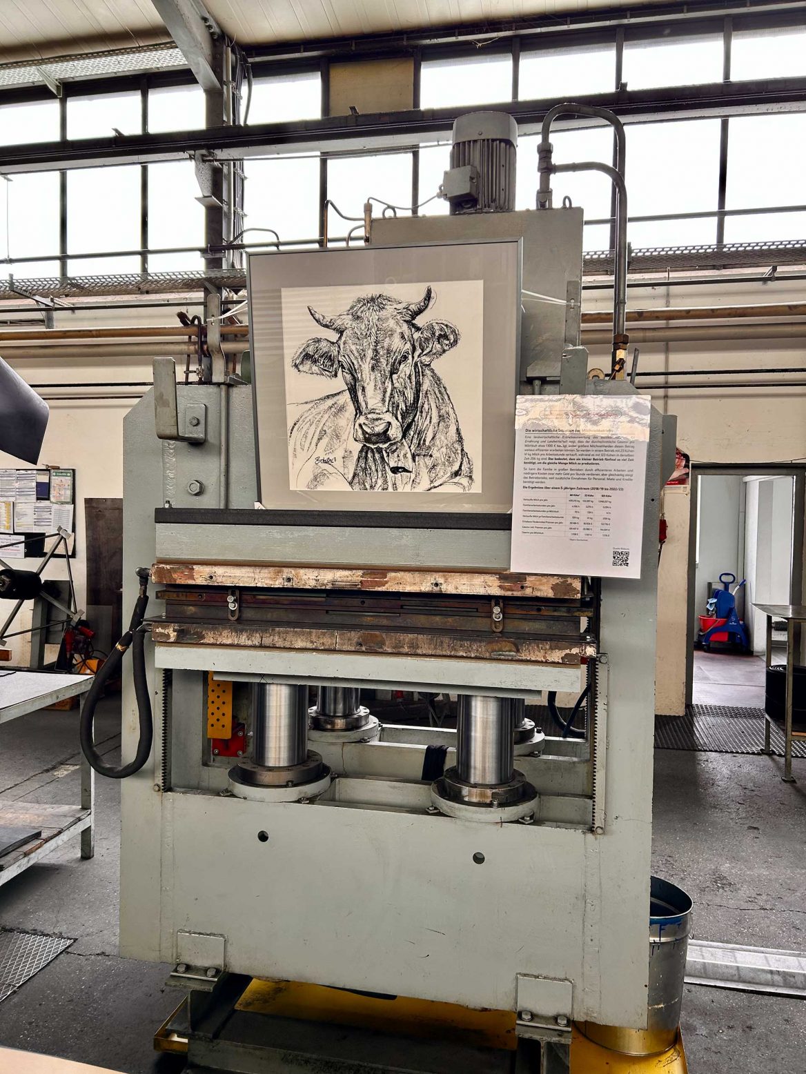 In einer Werkstatt hängt an eine großen Industriepresse eine gerahmte Kohlezeichnung einer Kuh. Daneben ist ein Blatt mit Informationen angebracht. Die Umgebung ist funktional mit anderen industriellen Geräten und hohen Fenstern im Hintergrund. Die Maschine ist massiv, mit sichtbaren hydraulischen Zylindern und mechanischen Komponenten.