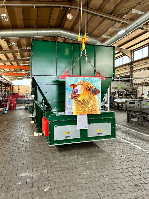 In einer großen Werkhalle hängt vor einem massiven grünen Sortiercontainer ein farbenfrohes Gemälde eines gelben Schafs, das von einem Deckenkran gehalten wird. Vor dem Gemälde hängt ein Blatt Papier mit Informationen zum Thema Schafe. Die Umgebung hat eine funktionale industrielle Ästhetik mit mehreren Maschinen und Ausrüstungsgegenständen.