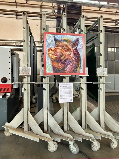 An mehreren großen Metallgestellen auf Rädern ist ein buntes Gemälde eines Wollschweins befestigt. Darunter ist ein Blatt mit Informationen über Schweine angebracht. Die Umgebung zeigt industrielle Ausrüstung und Rohre.