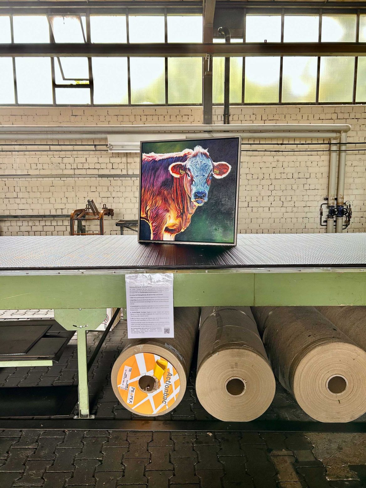 In einer Industriehalle steht auf einem großen grünen Förderband ein buntes Gemälde einer Kuh. Darunter ist ein Informationsblatt angebracht. Im Hintergrund sind hohe Fenster und eine Ziegelwand zu sehen. Unter der Metallfläche befinden sich riesige Papierrollen. Der Raum wirkt weitläufig und lichtdurchflutet.